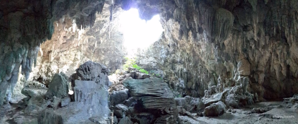 The interior of Bulwang Cave near Mabinay, Negros Oriental, Philippines, showing a vast limestone cavern with stalactites hanging from the ceiling, rugged rock formations on the cave floor, and a dramatic natural opening flooding the chamber with daylight.