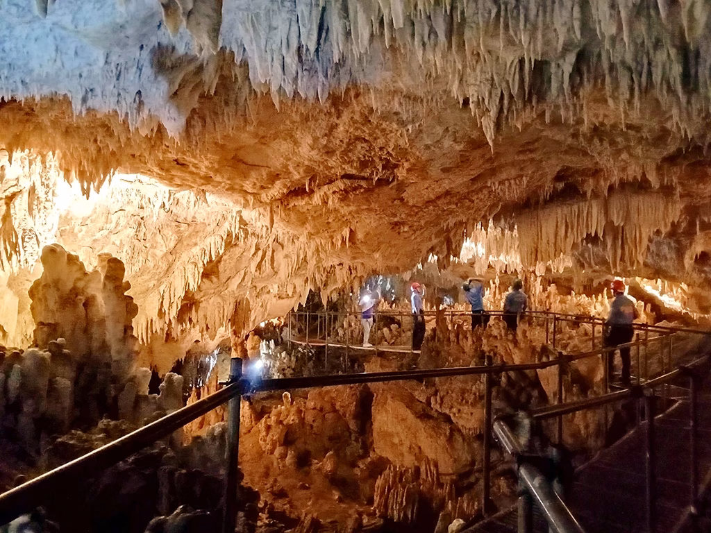 Tourists walk along a metal walkway inside the illuminated main chamber of Bulwang Cave in Mabinay, Negros Oriental, Philippines, surrounded by dense stalactite formations covering the ceiling and stalagmites rising from the cave floor, lit by warm amber lighting.