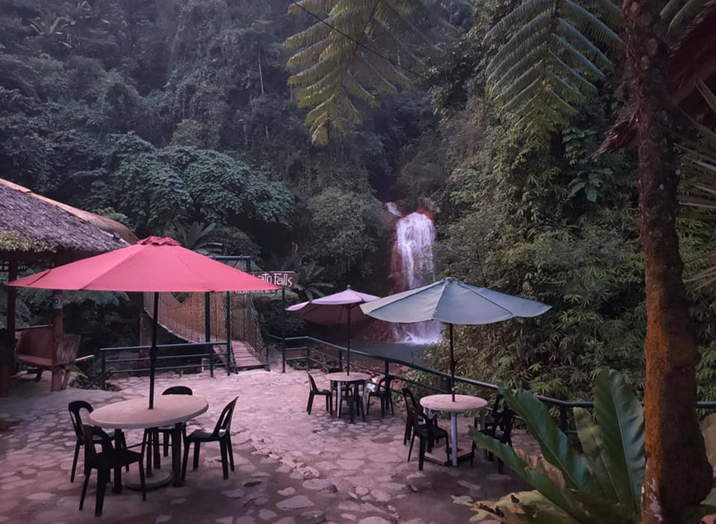 An open-air café with colorful umbrellas and plastic tables on a stone patio at the entrance to Casaroro Falls in Valencia, Negros Oriental, Philippines, with a suspension bridge, thatched nipa hut, and a waterfall visible through the surrounding jungle.