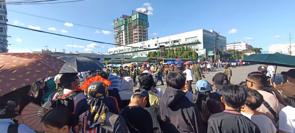 A massive crowd of Sinulog Festival 2025 devotees and spectators fills a Cebu City intersection, with umbrellas raised against the sun, security marshals in yellow vests managing the flow, and a festival dancer with a red headdress visible among the crowd.
