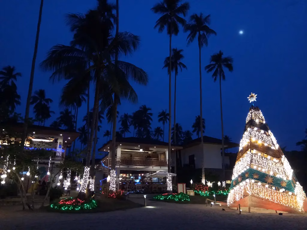 A large illuminated Christmas tree and festive light displays at a beachside resort in El Nido, Palawan, Philippines, at dusk, with tall coconut palms wrapped in white lights silhouetted against a deep blue evening sky and a single star visible above.
