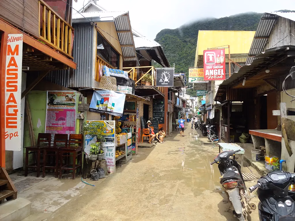 A narrow street in El Nido town, Palawan, Philippines, lined with small local businesses including a massage spa, fruit vendor, pawnshop, and money changer, with motorbikes parked along the muddy lane and a dramatic karst mountain rising into clouds at the end of the street.