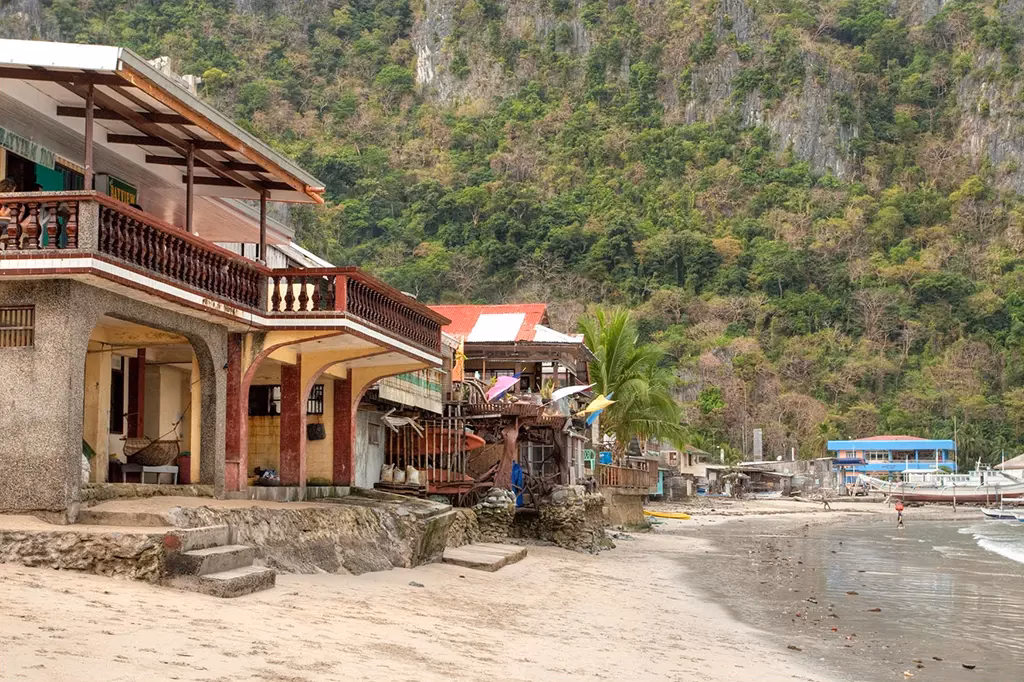 Shoreline restaurants and guesthouses lining the beach in El Nido town, Palawan, Philippines, with the buildings built directly onto the sand, a karst limestone cliff rising steeply behind them, and a quiet beach stretching toward Bangka boats in the distance.