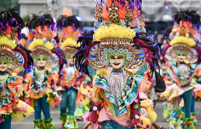 A close-up of a Sinulog Festival parade dancer in Cebu City, Philippines, wearing an ornate jeweled mask, towering feathered headdress, and a multicolored ruffled sequined costume, with a line of similarly costumed dancers visible in the background.