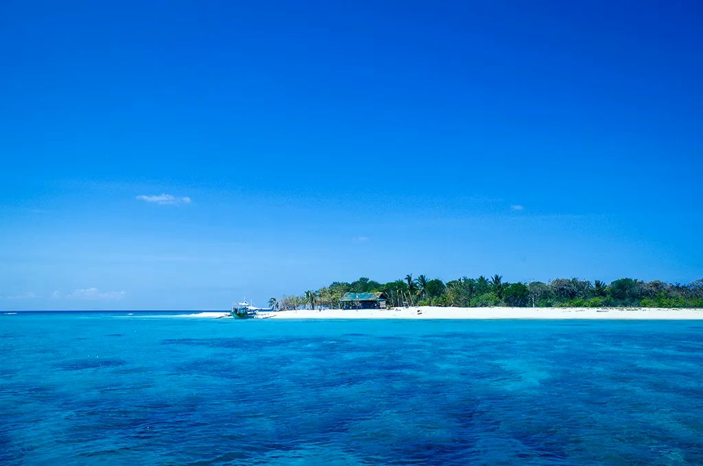 A small low-lying island with white sand beaches and coconut palms at Apo Reef Natural Park off Mindoro, Philippines, surrounded by vivid turquoise and cobalt blue water with coral reef clearly visible beneath the surface, and a bangka boat moored in the shallows under a deep blue sky.