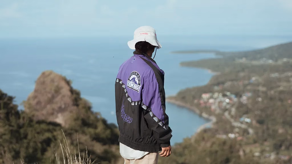 A visitor stands at a highland viewpoint looking out over a sweeping coastal bay with a town visible along the shoreline, a dramatic rocky peak in the foreground, and blue water stretching to the horizon at Cresta de Gallo Marine Protected Area in the Philippines.