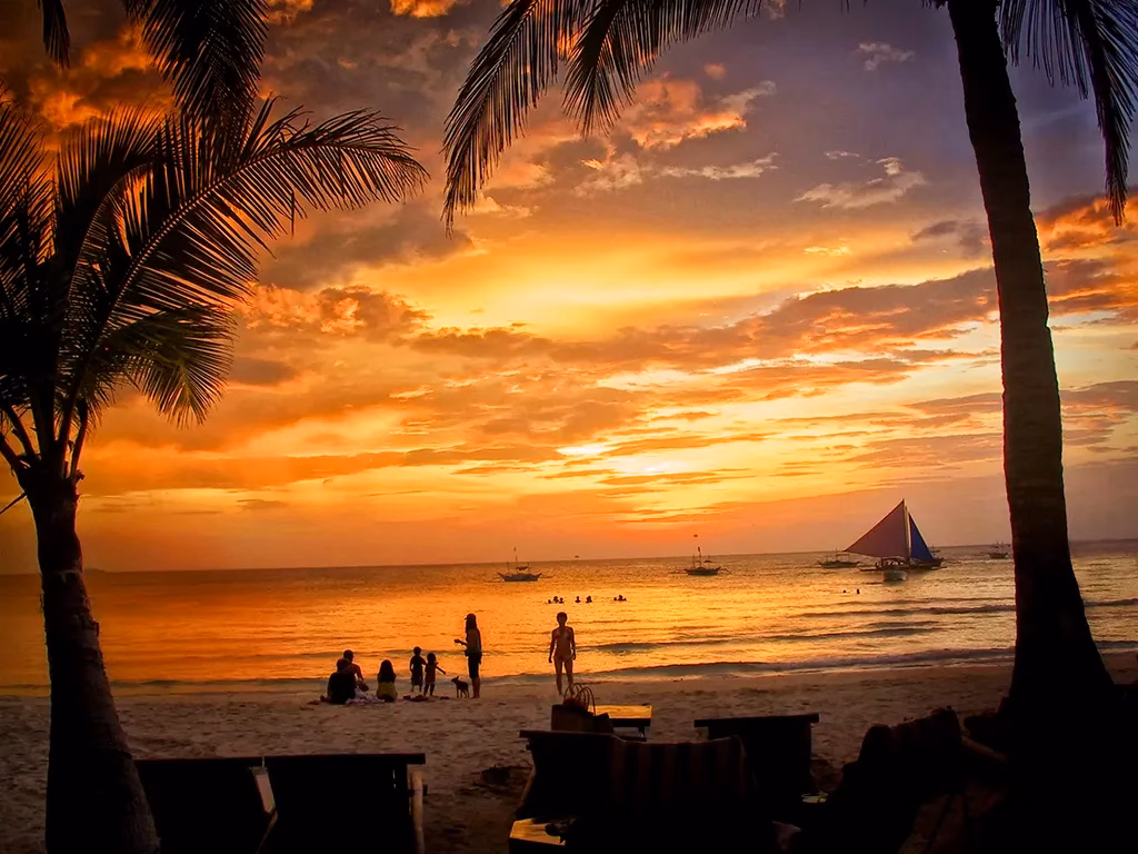 A dramatic fiery sunset over White Beach in Boracay, Philippines, with silhouetted coconut palms framing the scene, a traditional paraw sailboat on the glowing water, and silhouetted beachgoers and swimmers in the foreground under a sky blazing in orange and red.