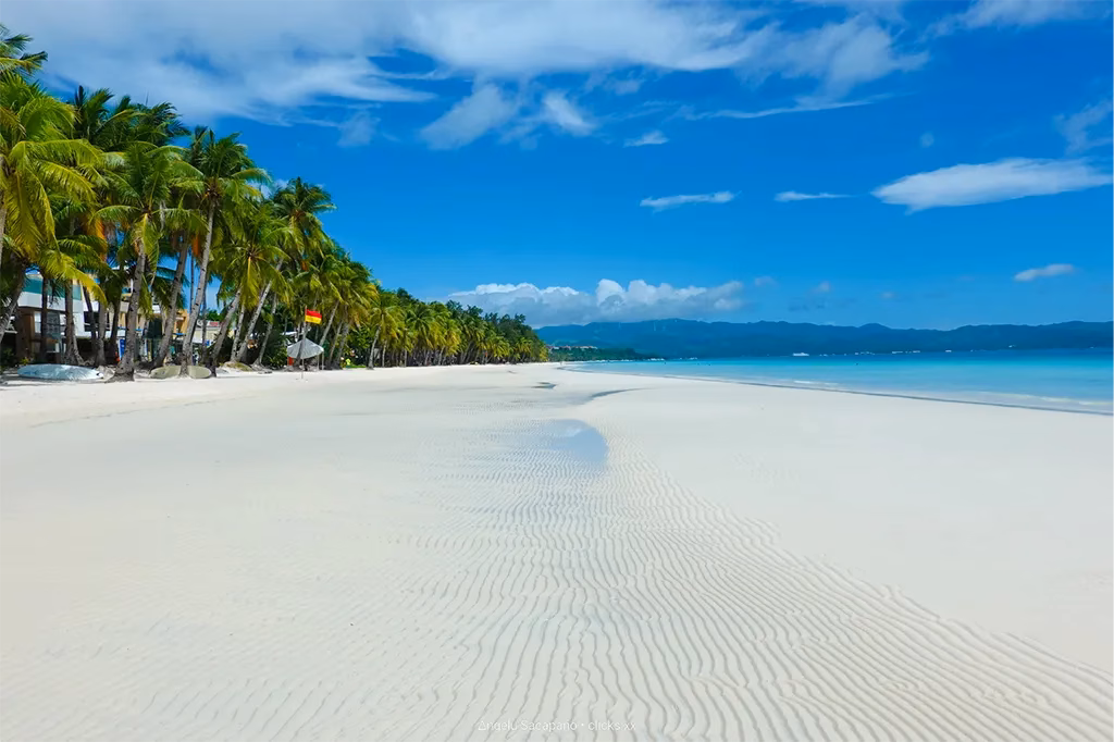 A wide expanse of powdery white sand on White Beach in Boracay, Aklan, Philippines, with coconut palms lining the shoreline, turquoise water stretching to the right, a lifeguard flag visible mid-beach, and the mountains of Panay Island visible across the strait under a blue sky with white clouds.