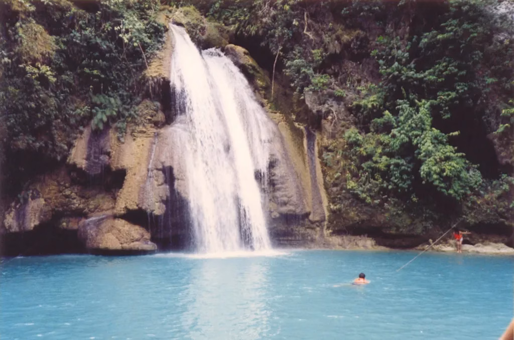 A swimmer in the vivid turquoise pool at the base of Kawasan Falls in Badian, Cebu, Philippines, with white water cascading down a tiered rock face surrounded by dense tropical jungle, and two visitors visible on the rocks to the right.