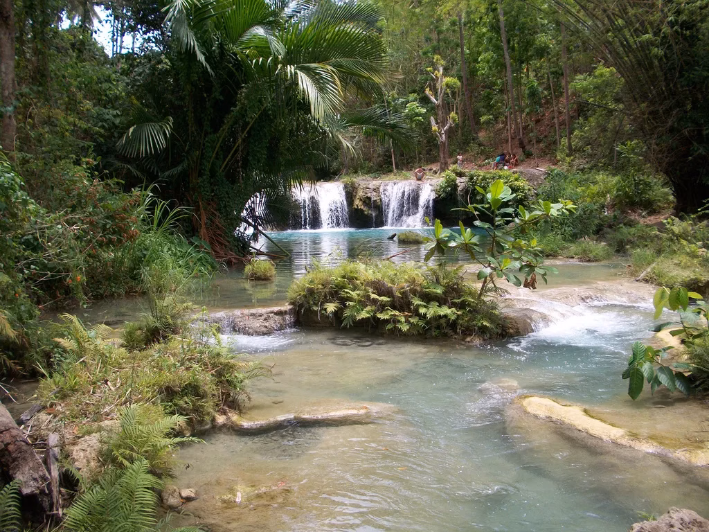 The multi-tiered cascades of Cambugahay Falls in Siquijor, Philippines, with turquoise water flowing through a series of connected pools surrounded by dense tropical jungle, ferns, and palm trees, with visitors visible on the upper tier in the background.