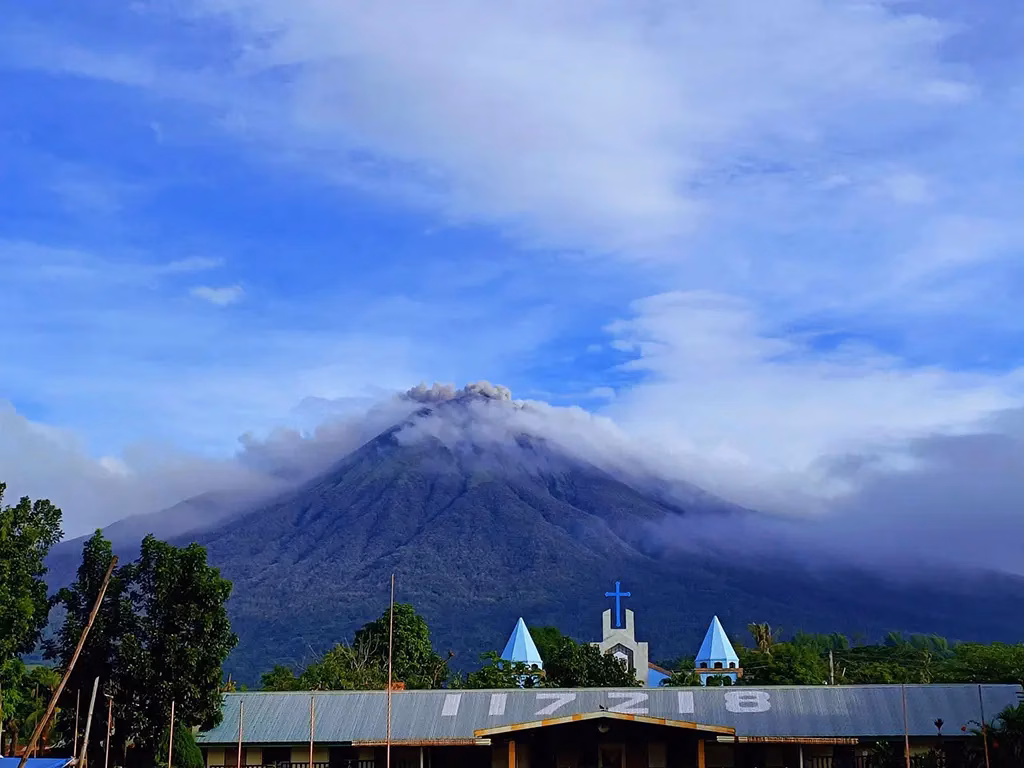 Kanlaon Volcano in Negros Oriental, Philippines, venting ash and steam from its summit while clouds wrap around its upper slopes, with a small church featuring a blue cross and cone-shaped turrets visible in the foreground.