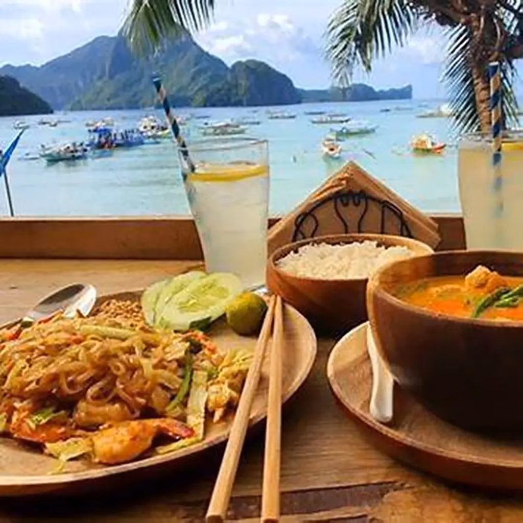 A beachfront restaurant meal in El Nido, Palawan, Philippines, featuring shrimp pancit noodles and an orange curry with rice served in wooden bowls with chopsticks and cold lemonades, with a view through the window of bangka boats on turquoise Bacuit Bay and karst islands in the background.