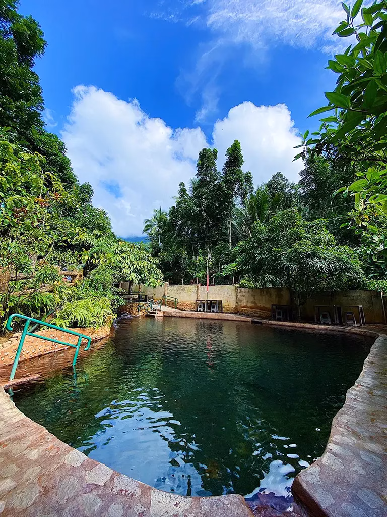 A geothermal hot spring pool with natural rock edges surrounded by dense tropical jungle at Red Rock Hot Spring in Valencia, Negros Oriental, Philippines, under a blue sky with white clouds, with metal entry railings and simple amenities visible along the far edge.