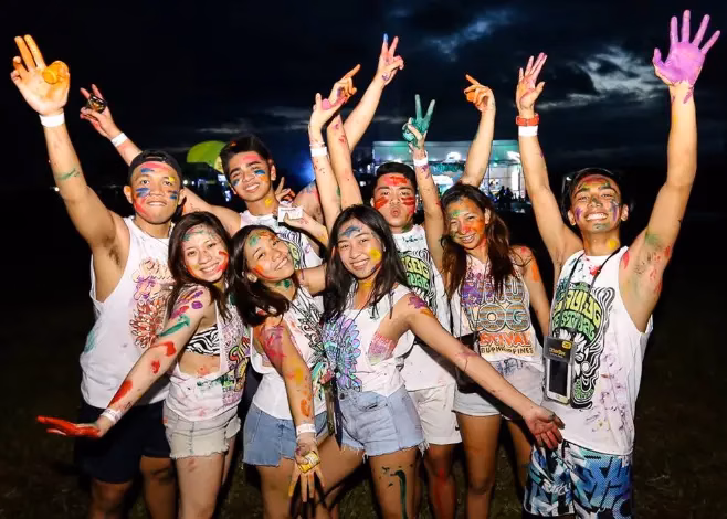 A group of young Sinulog Festival after-party attendees in Cebu City, Philippines, covered in colorful body paint with arms raised and smiling, wearing Sinulog festival shirts, celebrating at an outdoor night event after the main parade.