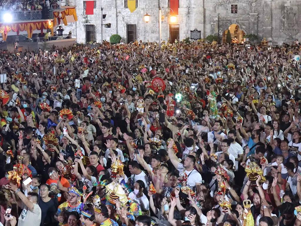 Thousands of devotees crowd the plaza of the Basilica Minore del Santo Niño de Cebu during the 2023 Sinulog Visperas Mass, arms raised and Santo Niño statues held aloft, with the illuminated historic stone facade of the basilica visible in the background.