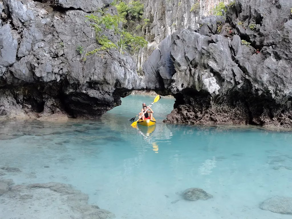 Two kayakers in a yellow kayak paddling through a narrow natural karst rock arch into the Small Lagoon on Miniloc Island in El Nido, Palawan, Philippines, with crystal clear turquoise water revealing the sandy bottom and coral below the surface.