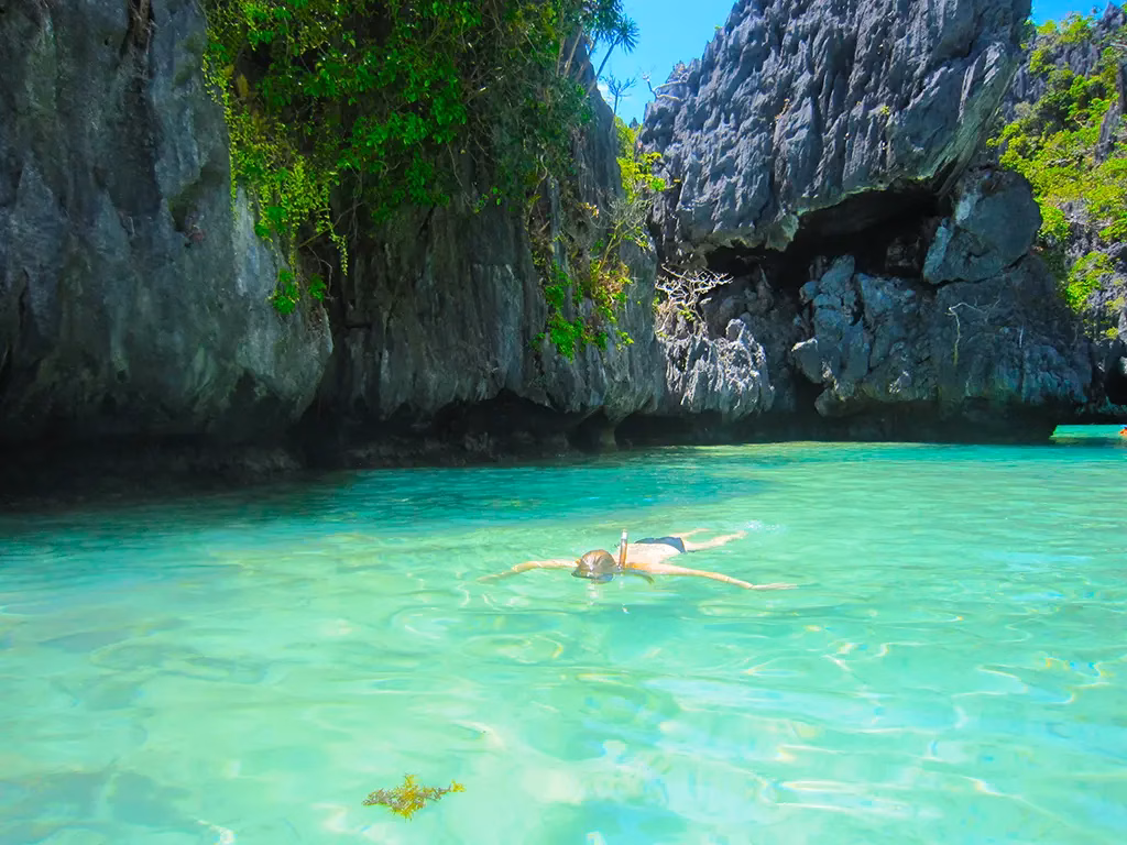 A snorkeler floating in the crystal clear shallow green water of the Green Lagoon in Bacuit Bay, El Nido, Palawan, Philippines, surrounded by towering karst limestone cliffs with a cave opening visible in the rock face and the sandy bottom clearly visible through the water.