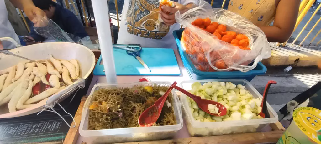 A Sinulog Festival street food vendor's stall in Cebu City, Philippines, displaying fresh lumpia wrappers, pancit noodles, chopped vegetables, and a bag of kwek-kwek orange battered quail eggs, with the vendor preparing orders for the festival crowd.
