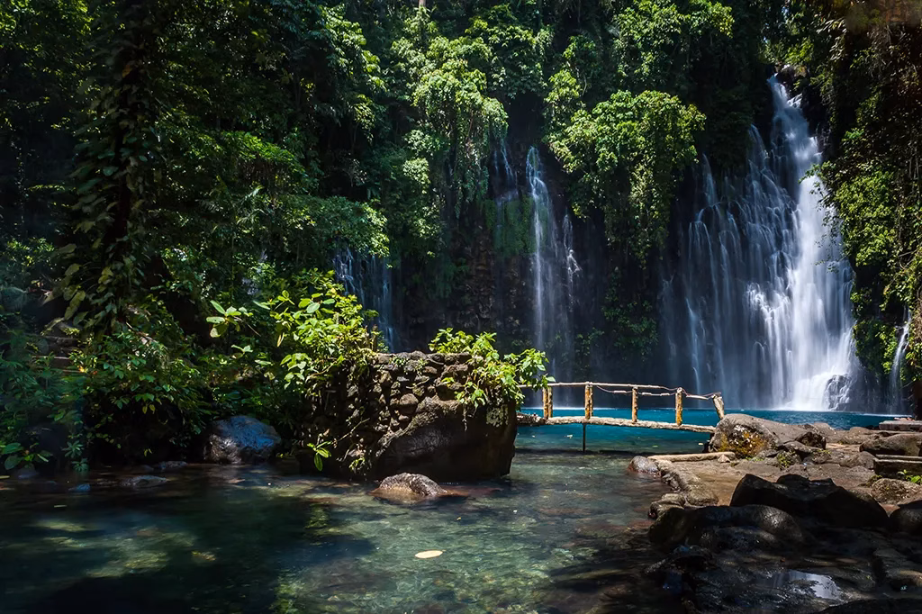 Tinago Falls in Iligan City, Lanao del Norte, Mindanao, Philippines, with multiple cascades of white water dropping into a vivid turquoise pool surrounded by dense jungle, a small bamboo footbridge visible in the middle ground.