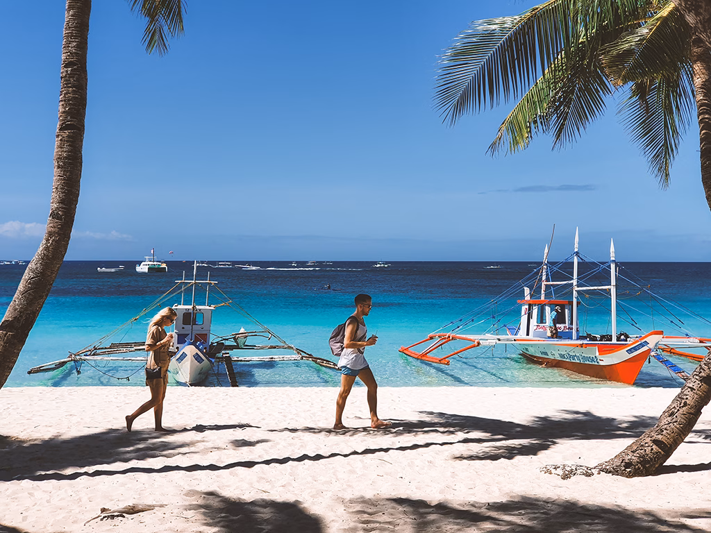 Two tourists walk along the powdery white sand of White Beach in Boracay, Malay, Aklan, Philippines, with traditional bangka outrigger boats beached in turquoise shallows and coconut palms framing the scene.