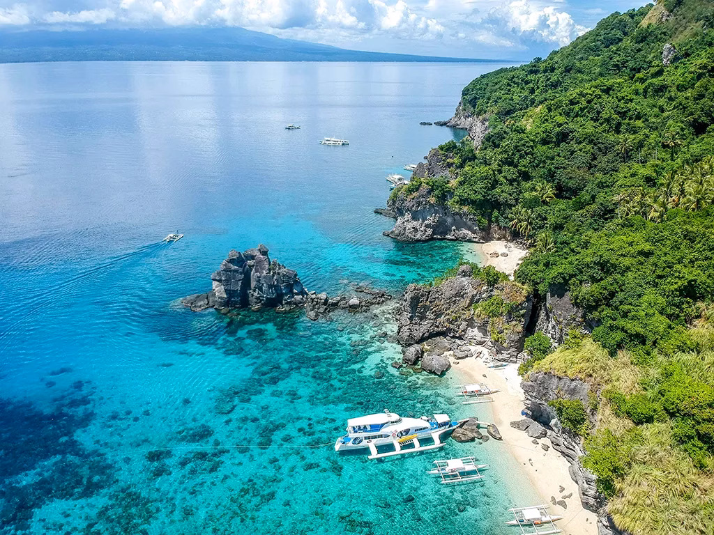 A boat on a white beach with trees and rugged rocks along the coastline. There are other boats offshore as well.